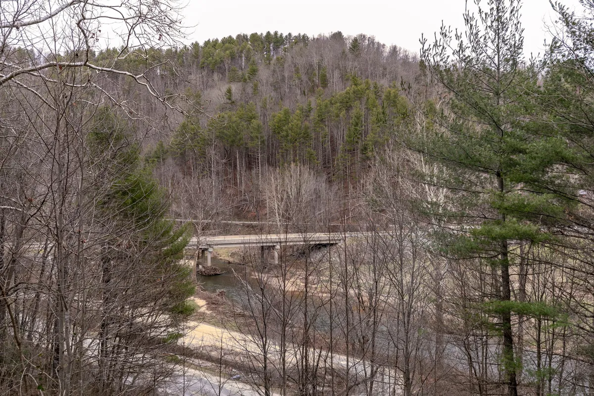 This view captures a serene landscape with a bridge over the river, dense forests, and the road winding alongside. It evokes tranquility and showcases the natural beauty waiting to be explored, hinting at activities like hiking, fishing, and nature walks.
