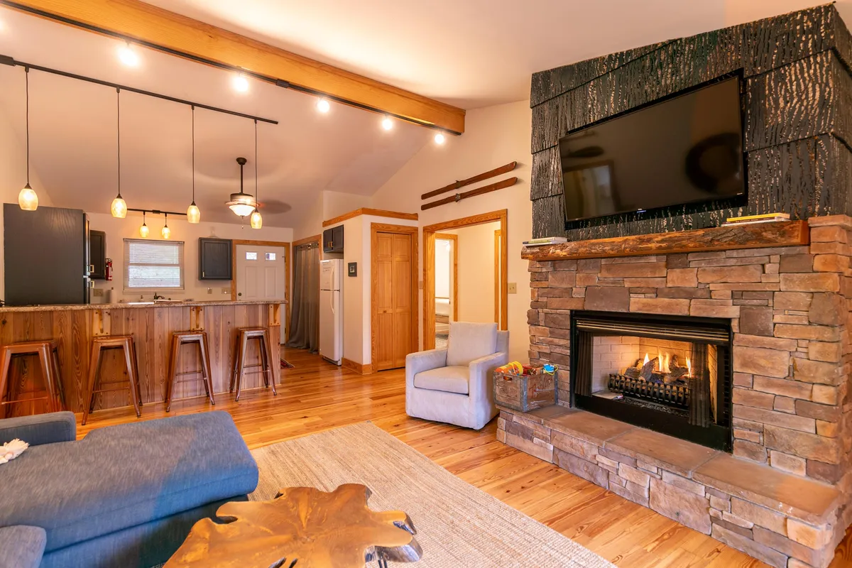 A spacious, open-concept living area with a prominent stone gas fireplace, comfortable seating arranged for conversation, and a glimpse into the adjacent kitchen bar. Warm wood finishes and ample lighting create a welcoming, cozy atmosphere perfect for relaxing or entertaining.
