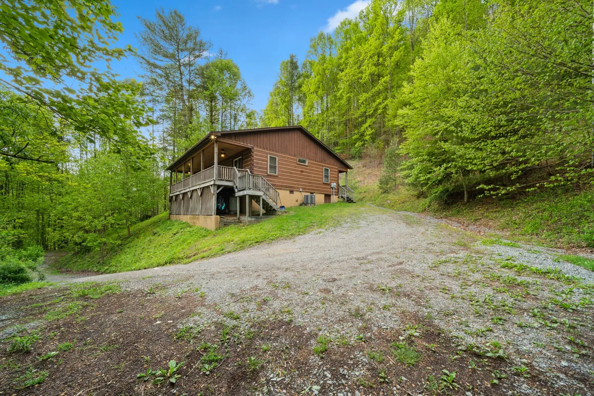 This image shows the exterior of a wooden house surrounded by lush greenery. The gravel driveway is clearly visible, winding up towards the property through the forested landscape. The scene looks peaceful and hints at the need for 4WD/AWD access due to the terrain.