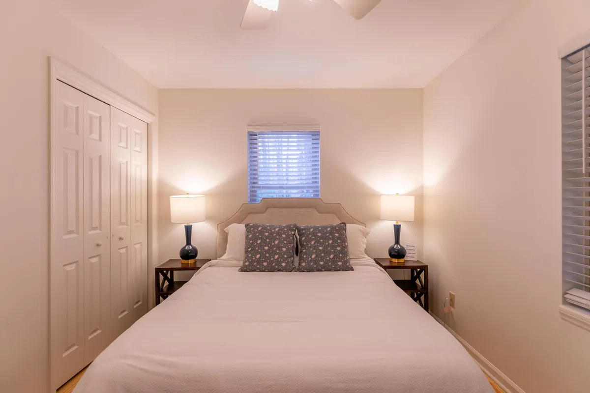 A serene queen bedroom featuring a comfortable bed, matching nightstands with soft-lit lamps, and a window letting in gentle natural light. The calm, neutral decor and tidy arrangement make the room inviting for restful nights or quiet moments.