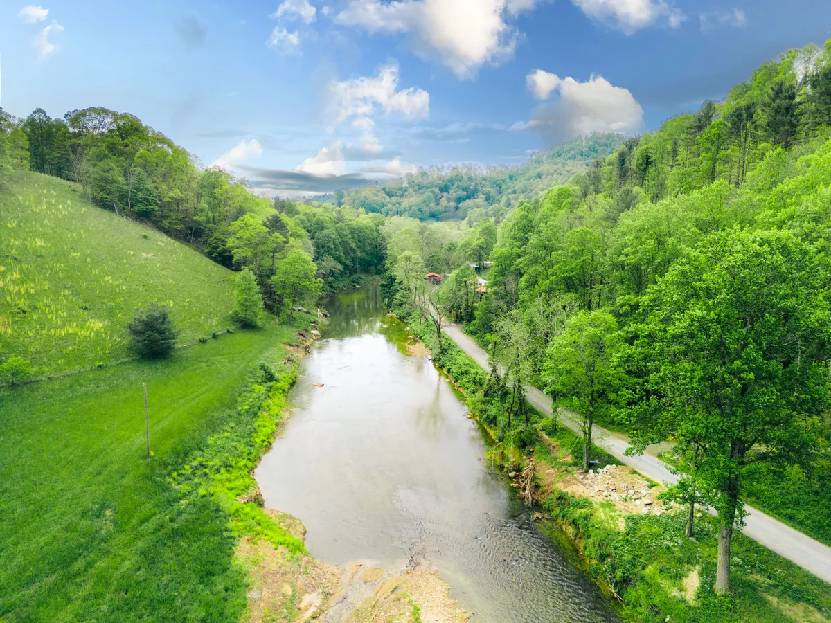 An expansive view of the river with lush greenery, rolling hills, and a peaceful roadway, showcasing the natural beauty and the proximity to the Watauga Gorge River Access. The scene is tranquil, with hints of outdoor adventure and relaxation.