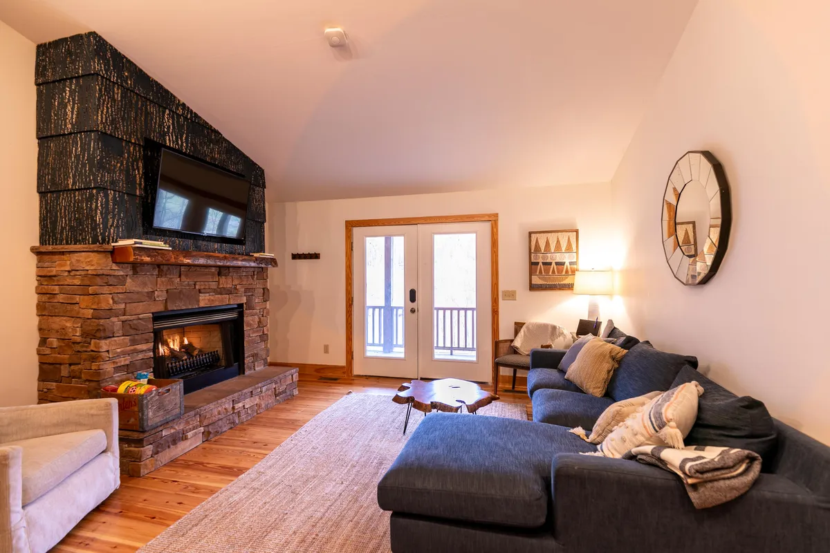 A cozy living room interior featuring a fireplace, plush sofa, coffee table, and double doors that lead outside. The space feels inviting and comfortable, suggesting a welcoming home base after outdoor adventures.