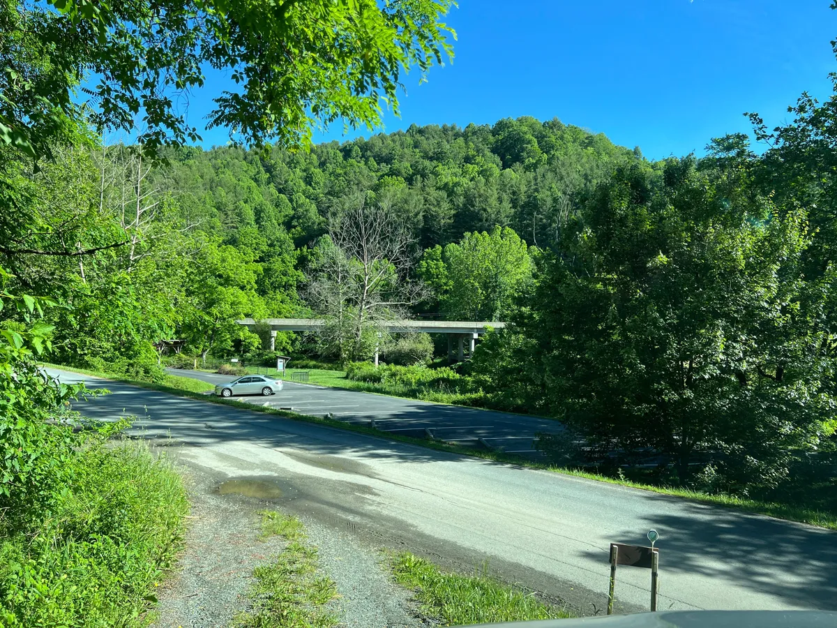 This exterior shot showcases the lush greenery, mountain backdrop, and a paved road with a parked car. The scene reflects the retreat's mountain setting and gives a sense of arrival and access to outdoor adventures.