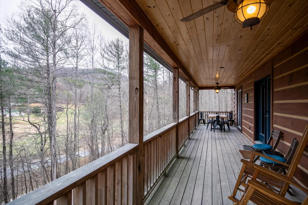 This image features a charming covered porch with a wooden railing, classic rocking chairs, a dining table, and the rustic exterior walls of the cabin. The porch looks out onto lush greenery, inviting guests to relax and enjoy the serene surroundings.