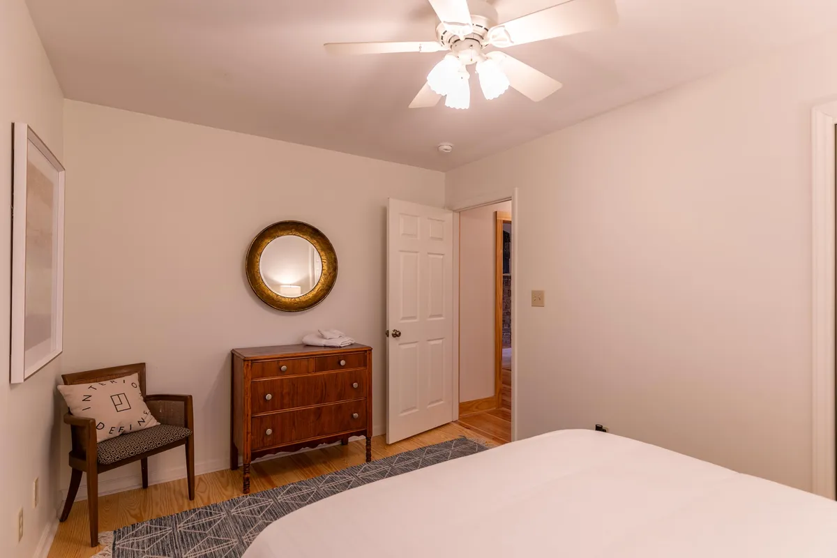 A calm and inviting bedroom with a bed dressed in white linens, a wooden dresser, a round mirror, and an accent chair, bathed in soft natural light. The arrangement radiates tranquility and comfort.