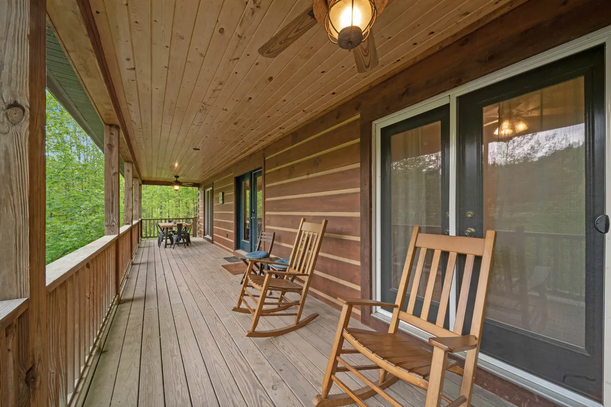 A pair of classic wooden rocking chairs are set on a spacious porch, with a table prepared for dining and lush greenery stretching into the background. The porch railings frame the peaceful view, suggesting a perfect spot for quiet reflection or enjoying the mountain air.