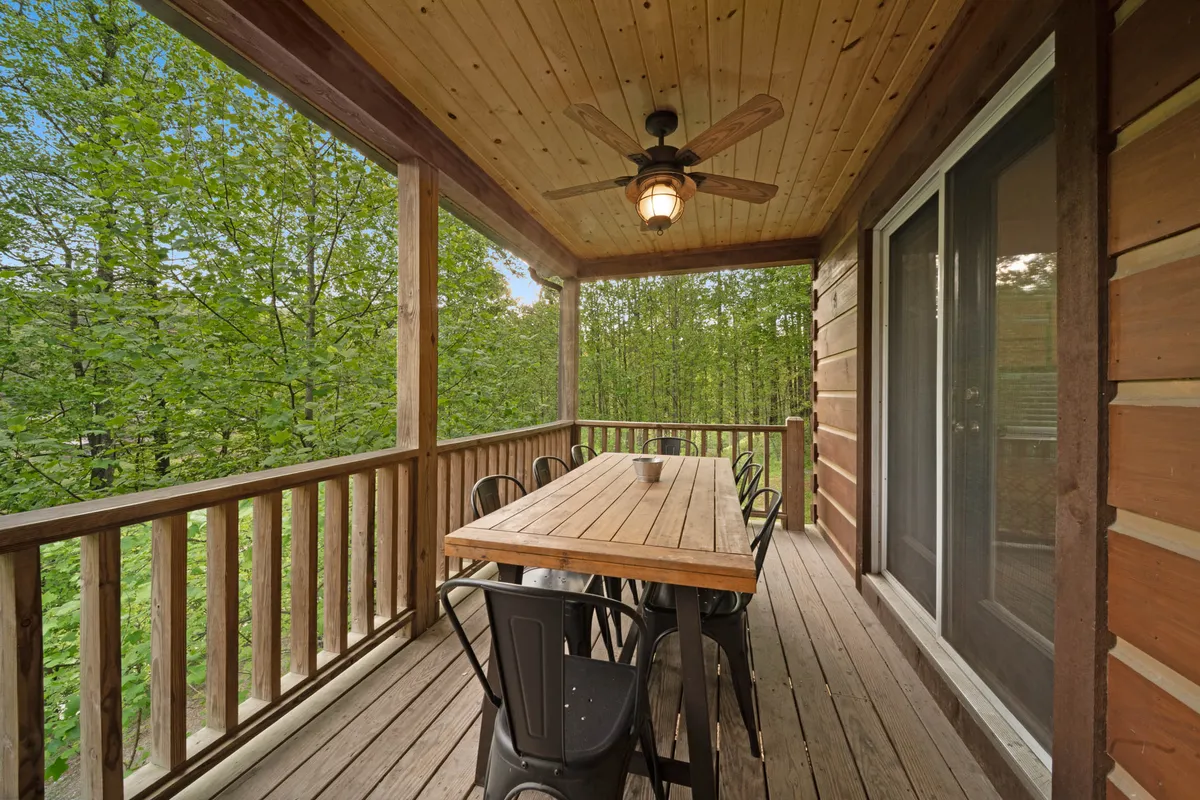 A welcoming outdoor dining area featuring a large wooden table, black metal chairs, and a ceiling fan, set against a backdrop of vibrant greenery. The arrangement suggests space for group meals, celebrations, or relaxed mornings surrounded by nature.