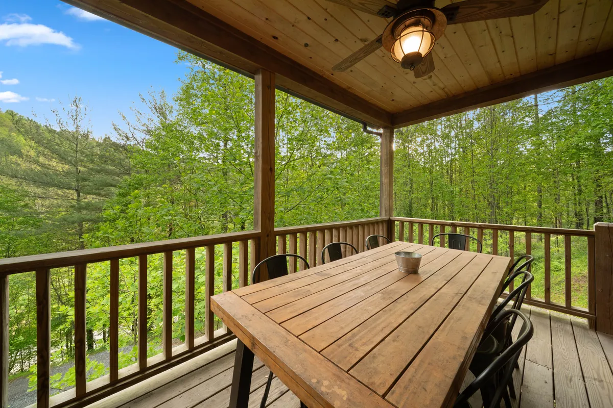 A welcoming outdoor dining area features a sturdy wooden table, several metal chairs, and is shaded by a ceiling fan with a light fixture. The space is surrounded by lush greenery, inviting guests to relax and dine al fresco while immersed in nature.