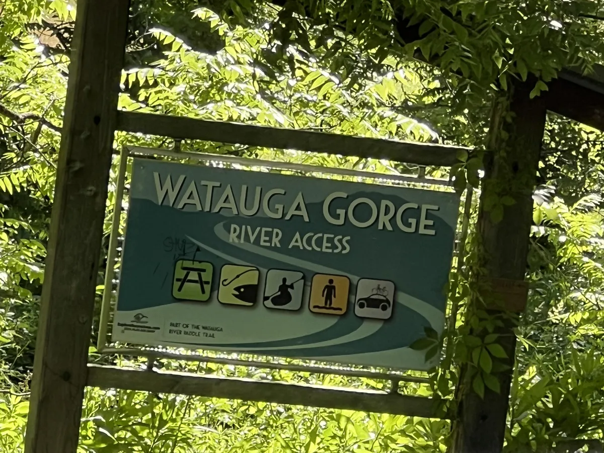 The image shows the Watauga Gorge sign surrounded by lush greenery, with a hint of the river nearby. Outdoor enthusiasts can easily identify the access point for fishing, kayaking, and other river activities. The scene suggests immediate proximity to adventure and highlights the natural landscape that makes the area special.