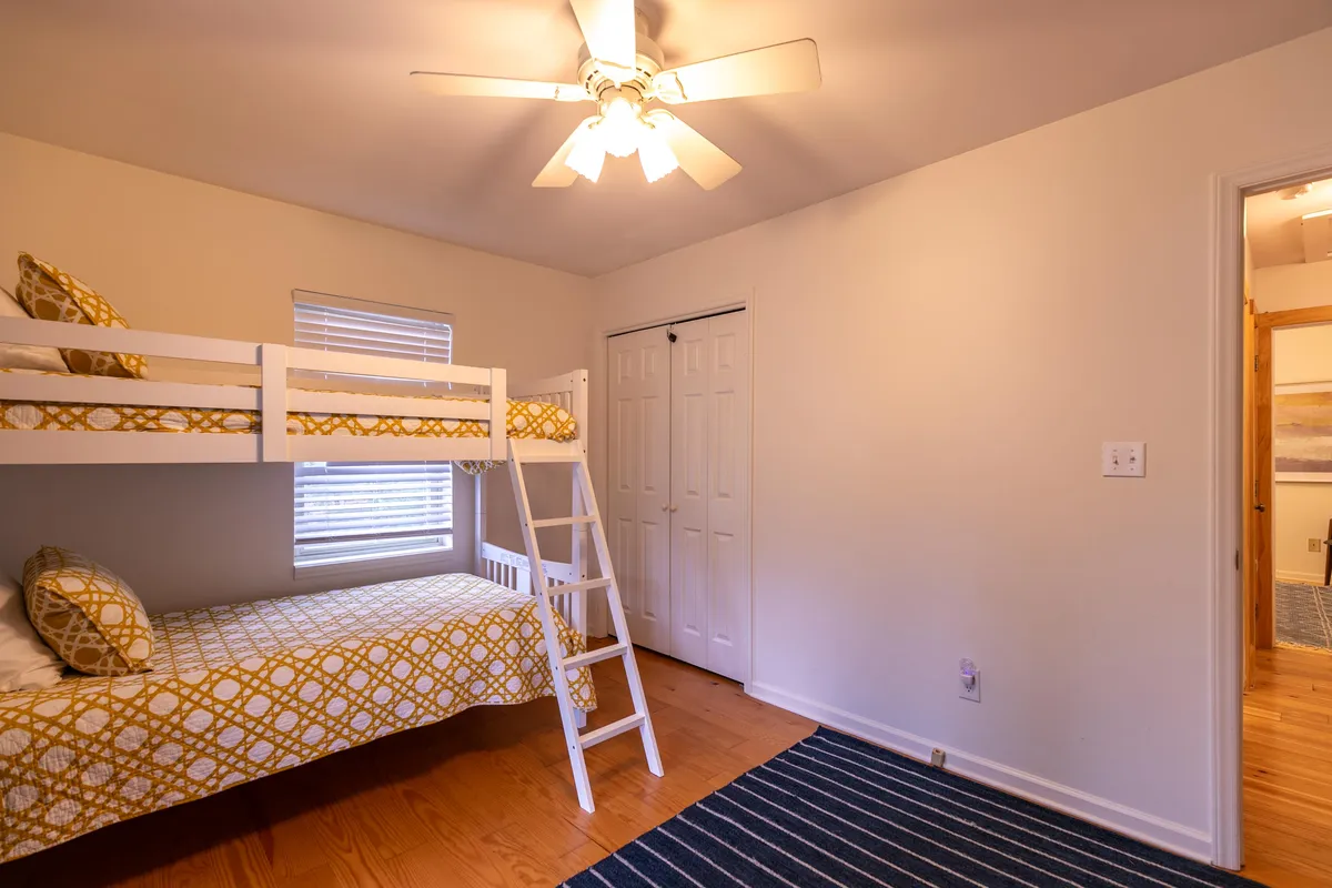 A cozy bunk room with a twin-over-twin bunk bed, a colorful striped rug, and a ceiling fan overhead. The room feels playful and welcoming, designed for children or siblings to share bedtime stories and laughter.