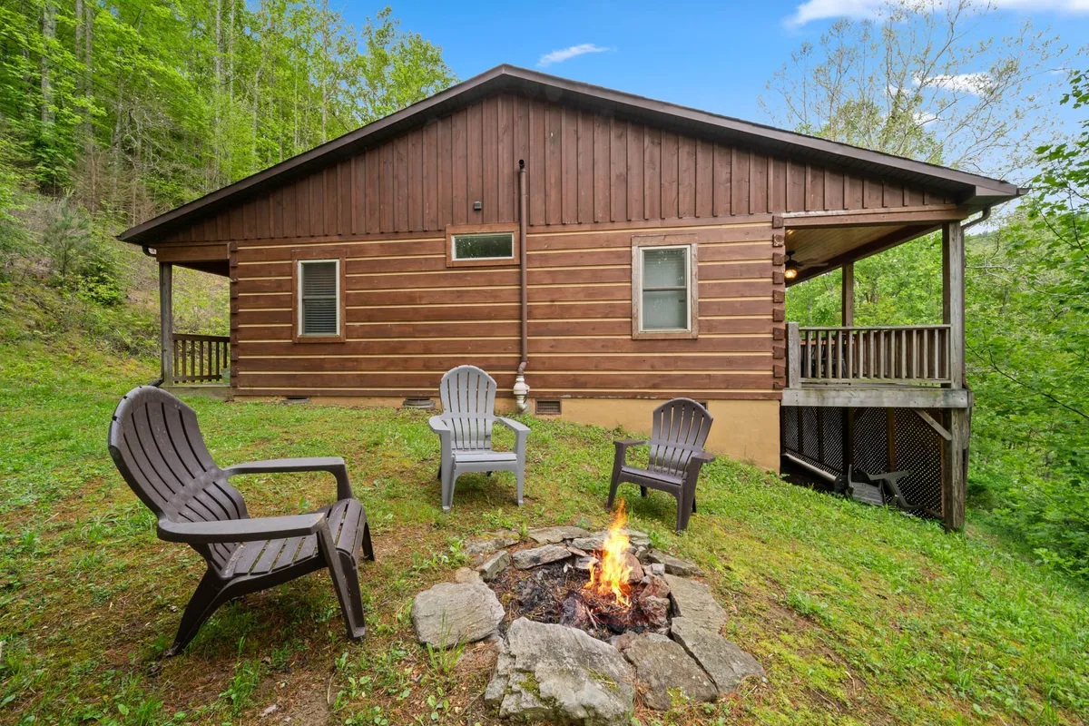 An exterior view of a cozy fire pit area surrounded by adirondack chairs and the wooden cabin in the background. Lush greenery frames the space, emphasizing the communal aspect of relaxing outdoors, sharing stories, or enjoying evenings around the fire with loved ones.